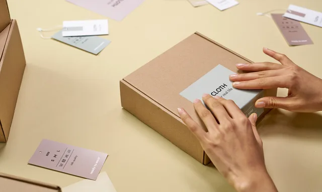 Hands carefully packing a cardboard box with multiple tags around, emphasizing sustainable packaging. Posizionamento di un brand DTC